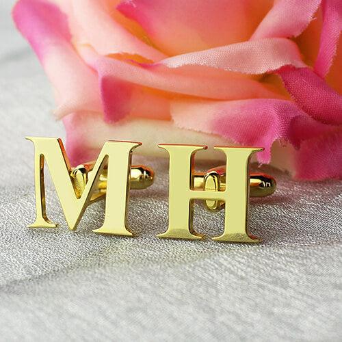 Gold initial cufflinks with the letters 'MH' displayed on a textured grey surface with a soft pink rose in the background.