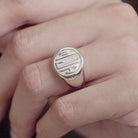 Fingers clasping an oval silver signet ring with a raised 'MJA' monogram, against a blurred background.