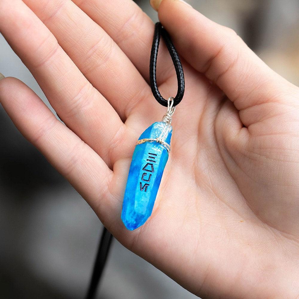 Hand holding a blue kyber crystal necklace with black cord and Aurebesh engraving.