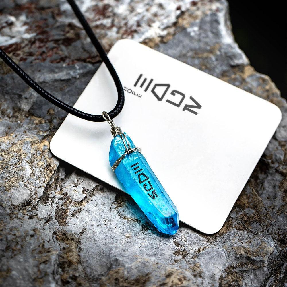 Blue kyber crystal pendant with Aurebesh on a card, against a rocky surface.