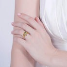 A woman's hand wearing a gold heart-shaped ring with colorful gemstones, resting gracefully on her opposite arm against a white dress.