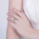 A woman's hand wearing a silver heart-shaped ring with colorful gemstones, resting gracefully on her opposite arm against a white dress.