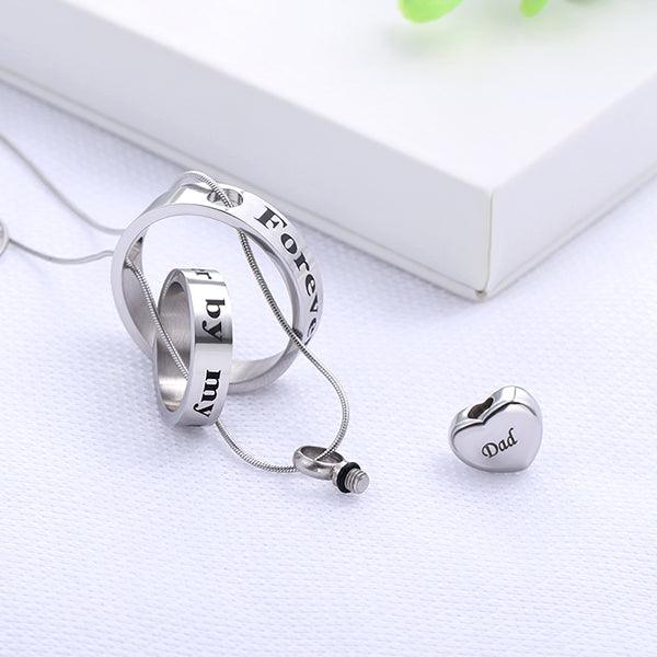 Silver memorial necklace with two engraved rings and separate heart pendant marked 'Dad', laid out beside a white box.