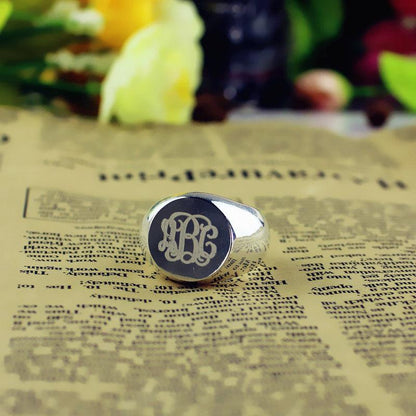 A polished silver signet ring with engraved ornate initials "LBC" rests on a vintage newspaper background with flowers blurred in the distance.