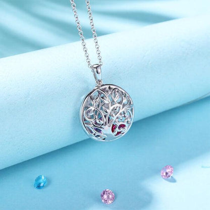 A silver necklace with a circular pendant featuring an intricate tree design, displayed on a light blue background with scattered colored gemstones.