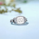 Silver ring with 'ASJ' monogram, diamond halo, and intricate side loops on a floral background.