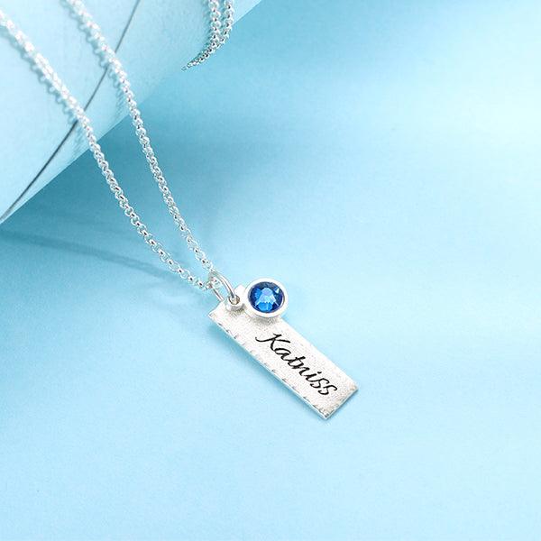 Elegant hammered personalized birthstone bar charm necklace in pure silver with the name "Katniss" and a blue gemstone, displayed on a blue background.