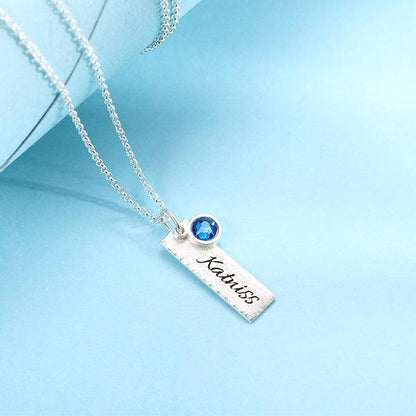 Elegant hammered personalized birthstone bar charm necklace in pure silver with the name "Katniss" and a blue gemstone, displayed on a blue background.