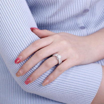 A woman wearing an elegant sterling silver engagement ring with a sparkling princess cut gemstone and shimmering accent stones, displayed on her hand against a light blue sweater.