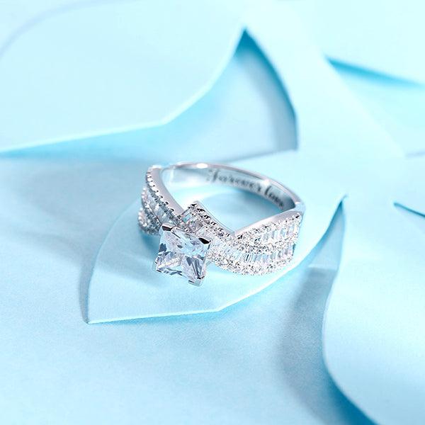 Elegant sterling silver engagement ring with a sparkling princess cut gemstone, shimmering accent stones, and personalized engraving, displayed on a light blue floral background.