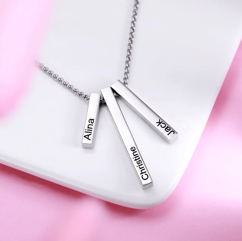 A close-up image of a silver necklace with three rectangular pendants engraved with the names "Alina," "Christine," and "Jack," placed on a white surface with a pink background.