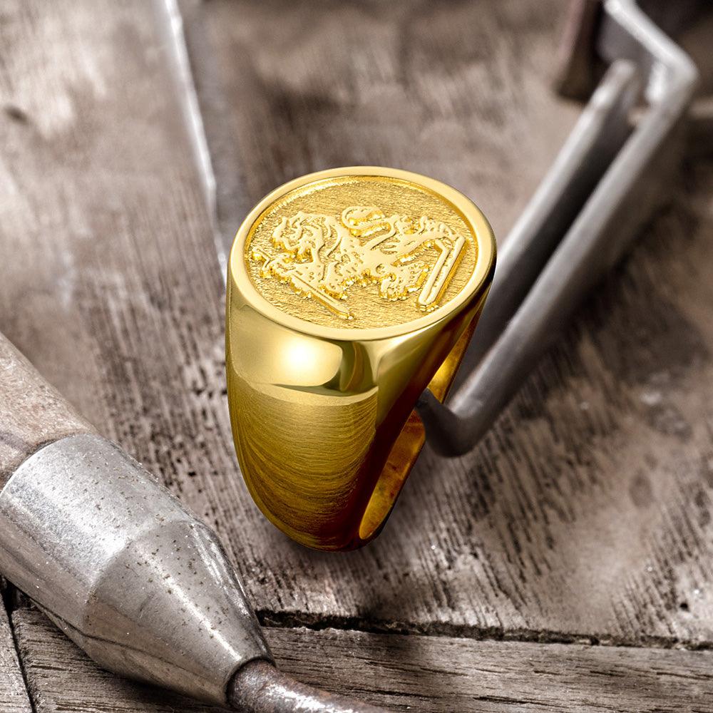 A golden signet ring with an intricate lion crest, resting on its side near jewelry-making tools on a wooden surface.