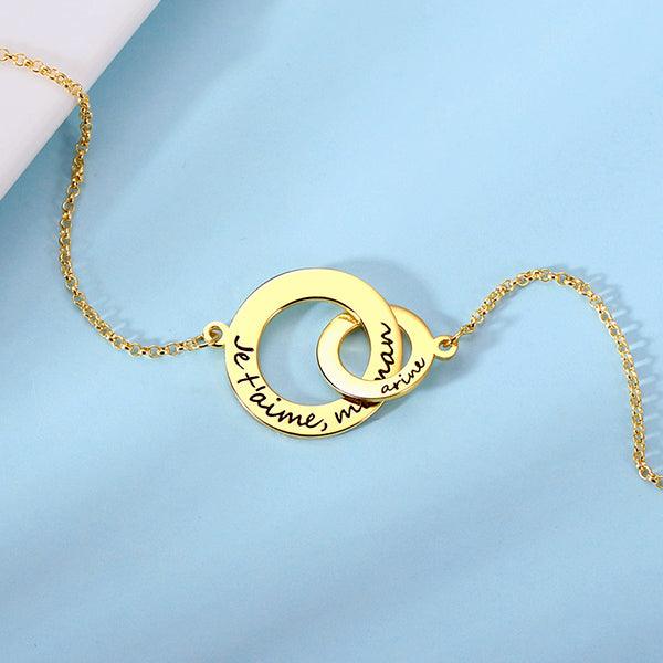 A gold necklace with interlocking circles. The larger circle has "Je t'aime, maman" engraved, and the smaller circle has the name "Aria" engraved.