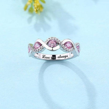 A silver ring with pink gemstones in a twisted design, surrounded by small clear stones, placed on a light blue background. The inside is engraved with "Love you always".