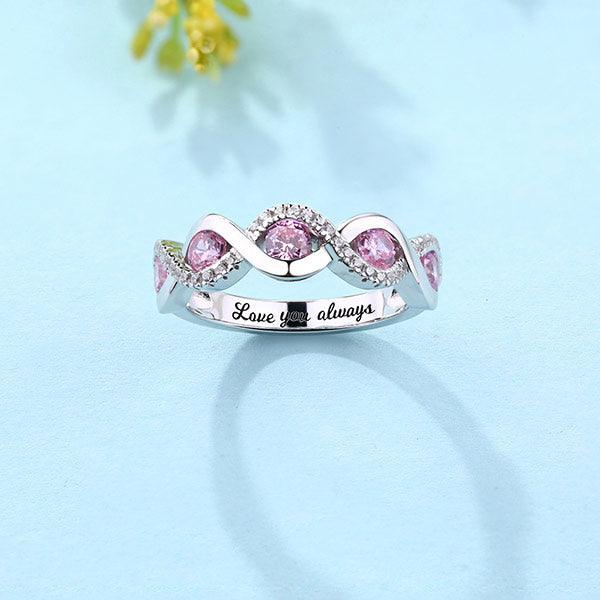 A silver ring with pink gemstones in a twisted design, surrounded by small clear stones, placed on a light blue background. The inside is engraved with "Love you always".