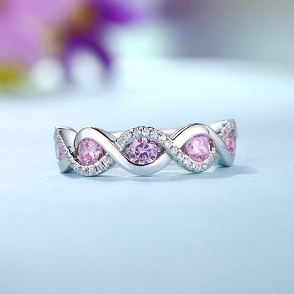 A silver ring with pink gemstones in a twisted design, surrounded by small clear stones, placed on a light blue surface with a blurred purple flower in the background.
