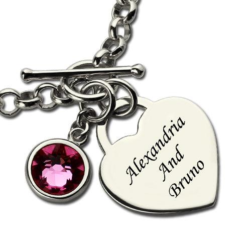 Close-up of a personalized heart charm bracelet with a pink birthstone and engraved names "Alexandria and Bruno," featuring a sterling silver toggle clasp.