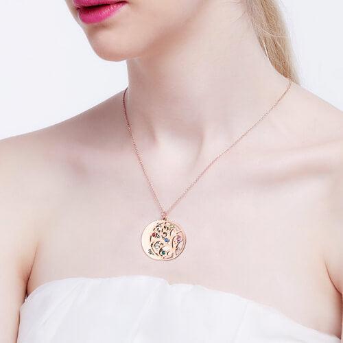 A woman wearing a rose gold double pendant necklace with a Tree of Life design featuring colorful gemstones. The pendant with engraved names is partially visible.