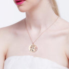 A woman wearing a rose gold double pendant necklace with a Tree of Life design featuring colorful gemstones. The pendant with engraved names is partially visible.