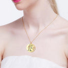 A woman wearing a gold double pendant necklace with a Tree of Life design featuring colorful gemstones. The pendant with engraved names is partially visible.