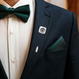 Man in navy suit with white shirt and green patterned bow tie, sporting a silver crest lapel pin.