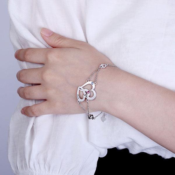 A person wearing a silver bracelet with two intertwined hearts, featuring the names Ella and Owen, and adorned with a red and a blue gemstone.