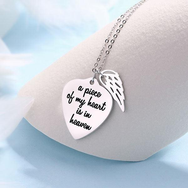 A silver necklace with a heart-shaped pendant engraved with "a piece of my heart is in heaven" and an angel wing charm, displayed on a light blue background.