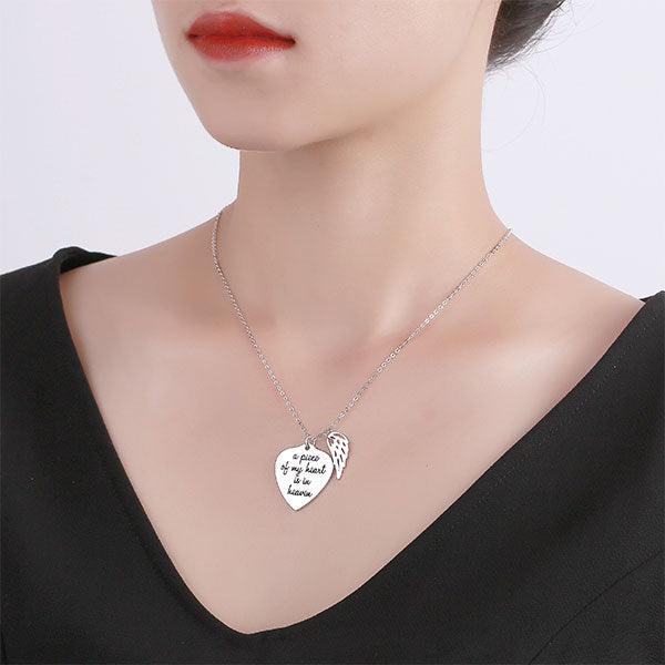 A woman wearing a silver necklace with a heart-shaped pendant engraved with "a piece of my heart is in heaven" and an angel wing charm.