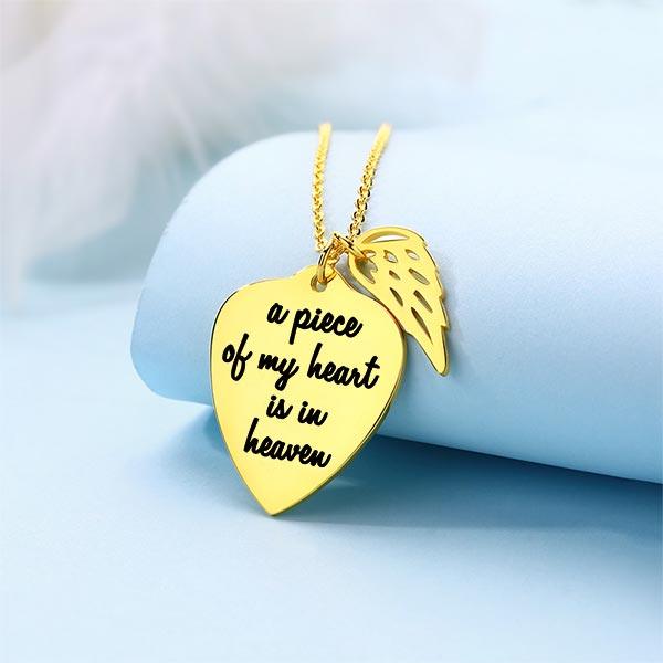 A gold necklace with a heart-shaped pendant engraved with "a piece of my heart is in heaven" and an angel wing charm, displayed on a light blue background.
