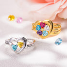 Two personalized heart-shaped rings with engraved names and colorful birthstones, one in silver and one in gold, on a soft fabric background with scattered gemstones.