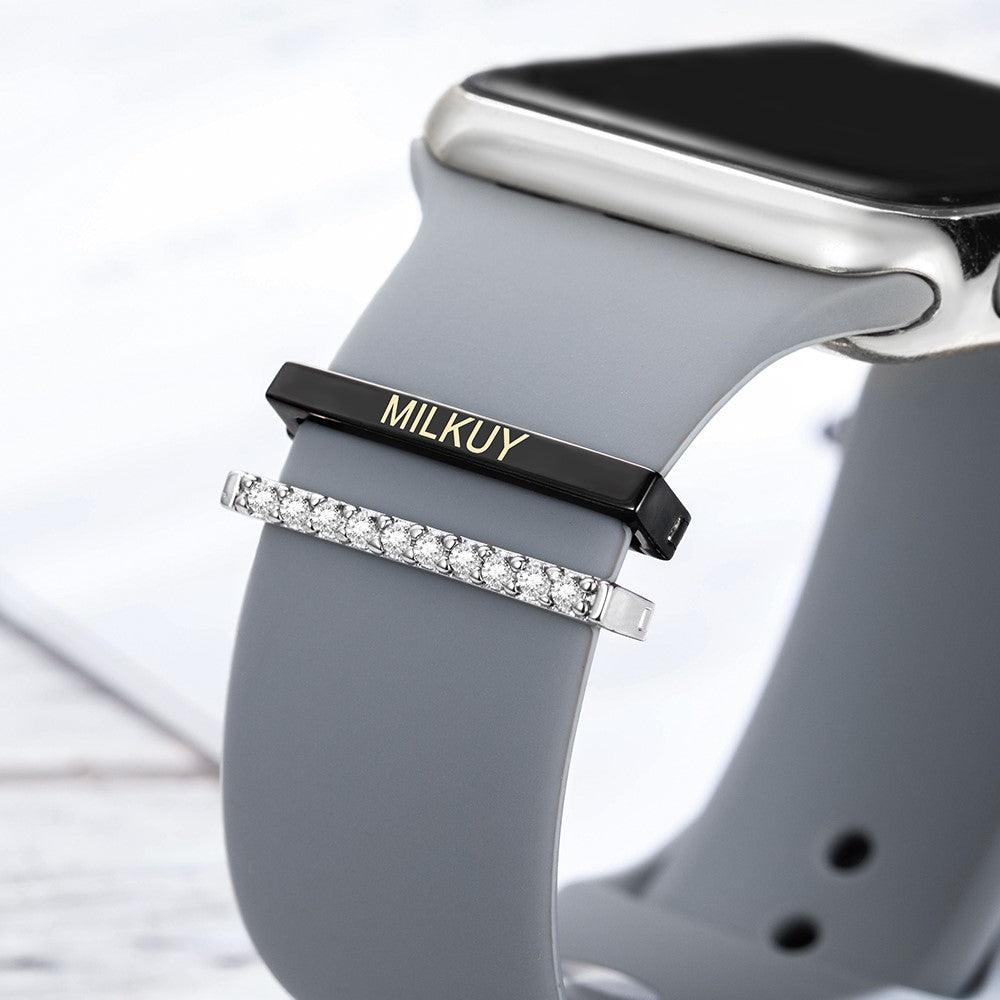 Close-up of an Apple Watch with a gray band, featuring personalized black and silver charms with the name "Milroy" and diamond-like accents.