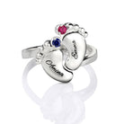 A silver ring with two engraved baby feet, featuring the names "Amina" and "Emma" with pink and blue gemstones, reflecting the inscription "Always in my heart."