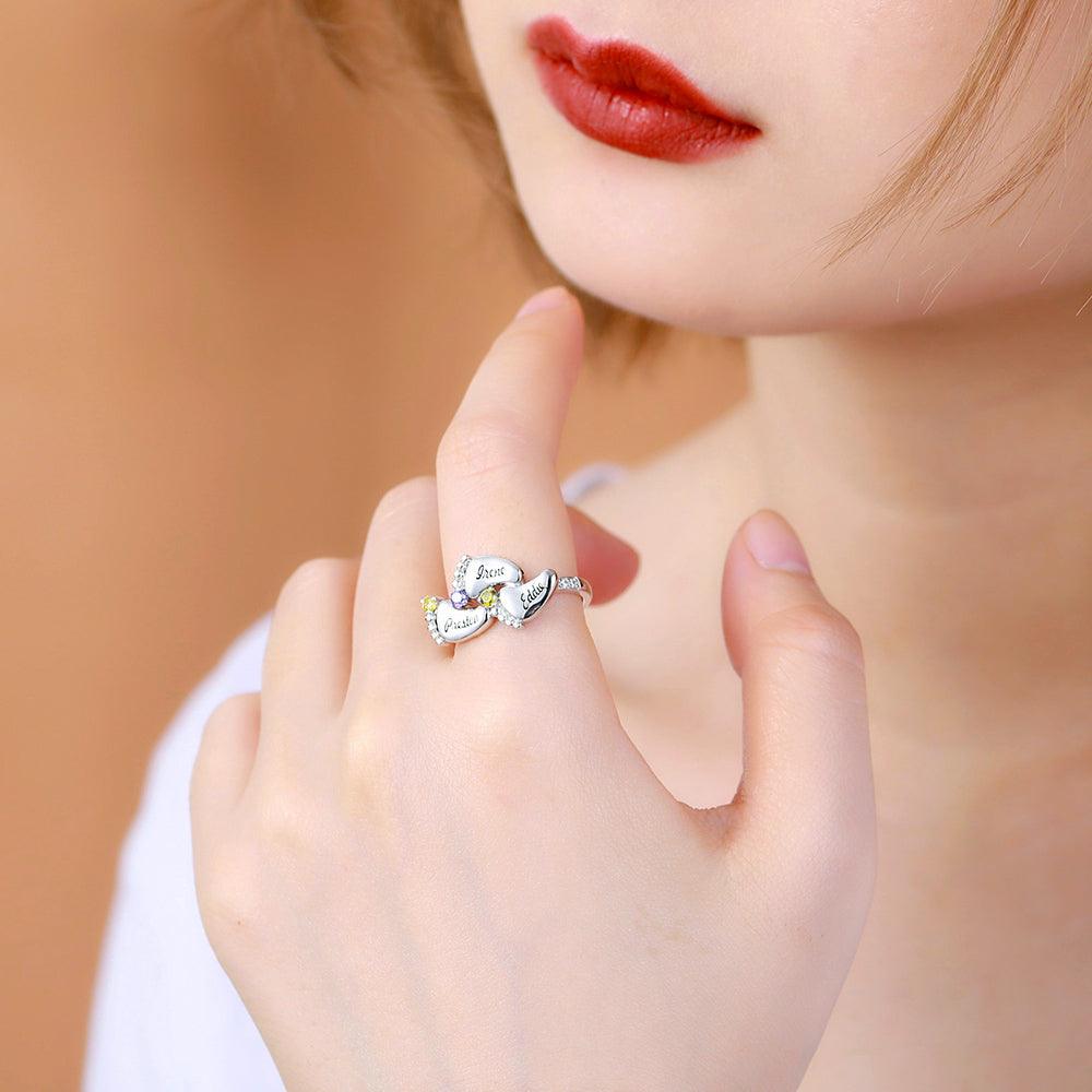 A woman wearing a silver ring with four engraved baby feet names and colorful gemstones, symbolizing "Always in my heart." She has short hair and red lipstick.