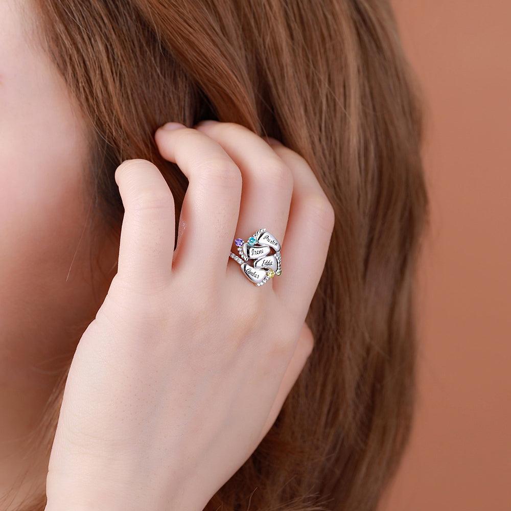 A woman wearing a silver ring with four engraved baby feet names and colorful gemstones, symbolizing "Always in my heart." She has light brown hair.