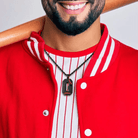 A man wearing a red baseball jersey and holding a bat over his shoulder, showcasing a black pendant shaped like the letter "G" with red baseball stitching details and the name "Gordon" engraved on it, on a black rope chain.