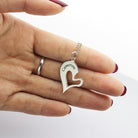 A hand holding a silver heart-shaped pendant with a cutout design and "Cameron" engraved on it, attached to a silver chain. The nails are painted red.