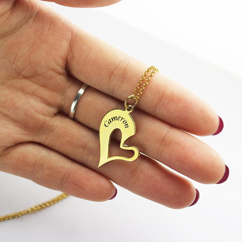 A hand holding a gold heart-shaped pendant with a cutout design and "Cameron" engraved on it, attached to a gold chain. The nails are painted red.