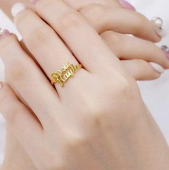 Close-up of a hand wearing a gold ring with cut-out name lettering in cursive. The background shows another hand with painted nails, slightly out of focus.