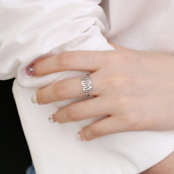 Close-up of a hand wearing a silver ring with cut-out name lettering in cursive. The hand is resting on a white fabric, with painted nails adorned with small gems.