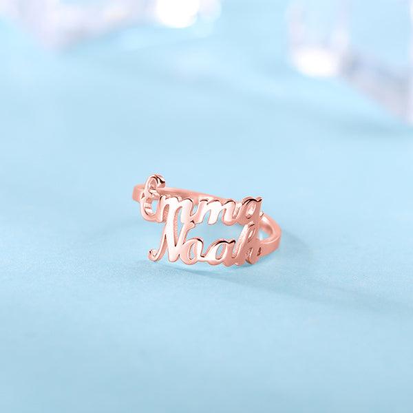 Rose gold ring with cut-out names "Emma" and "Noah" in cursive, placed on a light blue surface with a soft-focus background.