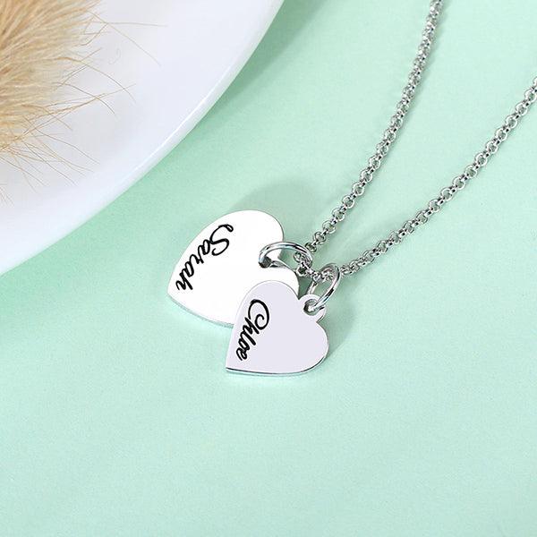 A silver necklace with two heart-shaped pendants, one engraved with "Sarah" and the other with "Chloe," displayed on a light green background near a fluffy item.