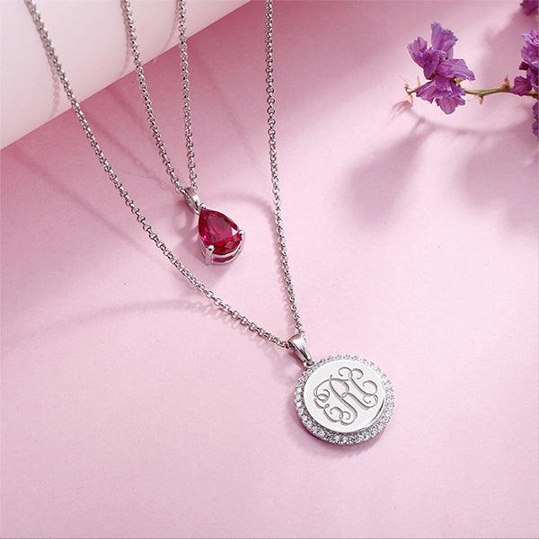 Double-layered necklace in sterling silver with a teardrop birthstone pendant and CZ monogram pendant, displayed on a pink background with purple flowers.