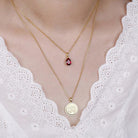 Double-layered necklace in gold with a teardrop birthstone pendant and CZ monogram pendant, worn on a woman's neck with a white lace top.