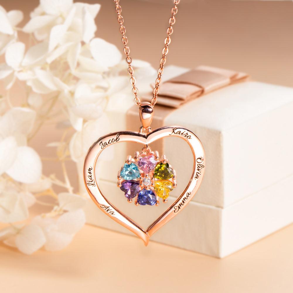 A heart-shaped rose gold necklace with colorful birthstones and engraved names, beautifully displayed on a gift box with white flowers in the background.