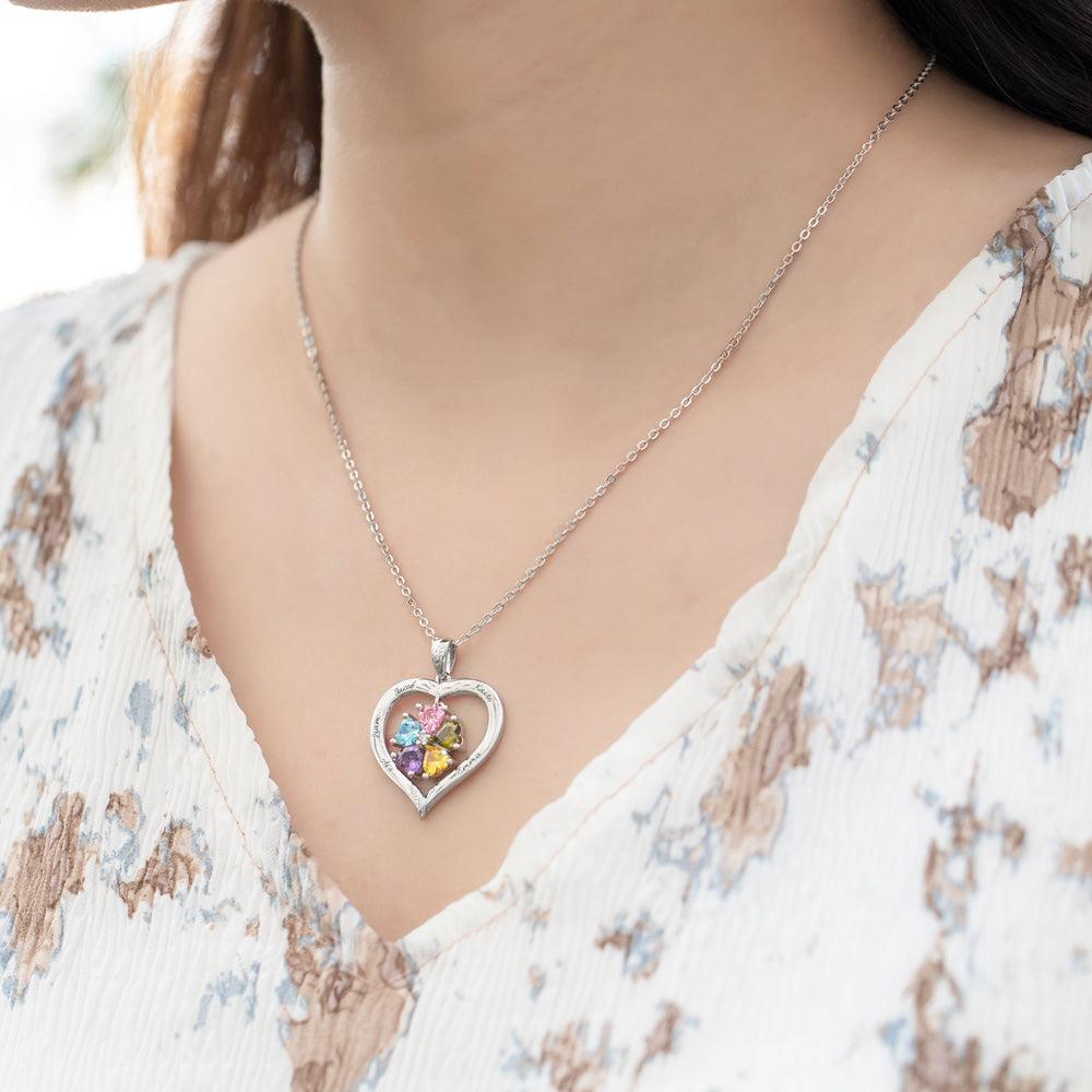 A woman wearing a heart-shaped silver necklace with colorful birthstones and engraved names, paired with a light floral blouse.