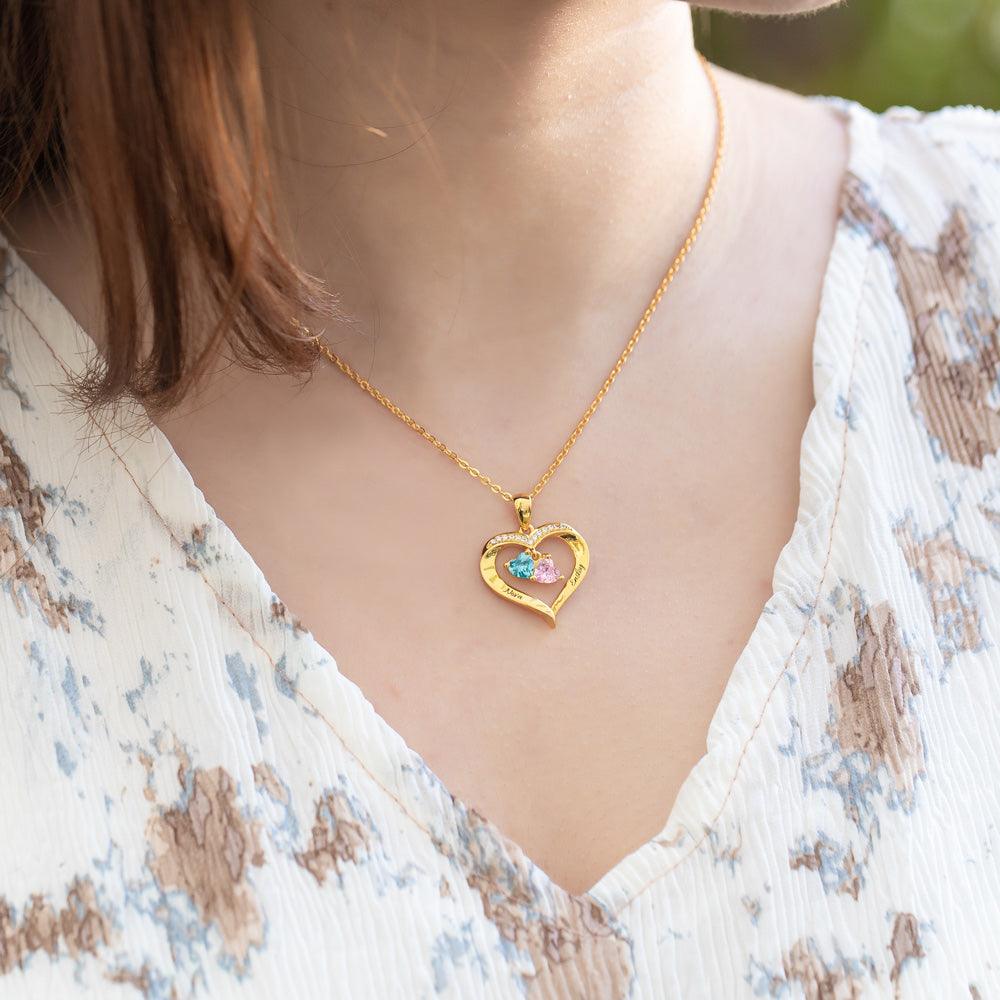 A woman wearing a heart-shaped gold necklace with colorful birthstones and engraved names, paired with a light floral blouse.