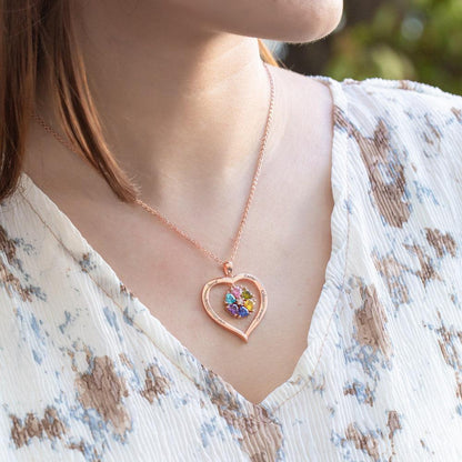 A woman wearing a heart-shaped rose gold necklace with colorful birthstones and engraved names, paired with a light floral blouse.