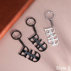 Three "DAD" keychains, two in black and one in silver, with personalized names, on a dual-tone background.