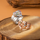 Two rings on a wooden surface: a silver one engraved with "Luke, Nathan, Liam, Ross" and a gold one with "Selena, Julian, Diane," both with floral designs and colorful gems.