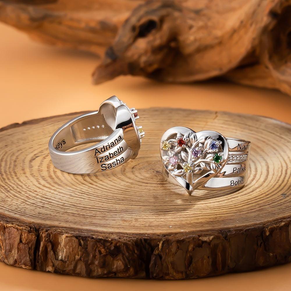 Two silver rings on a wooden surface: one engraved with "Adriana, Izabeth, Sasha" and the other with "Nixon, Parks, Bohdi," both featuring heart and floral designs with colorful gems.